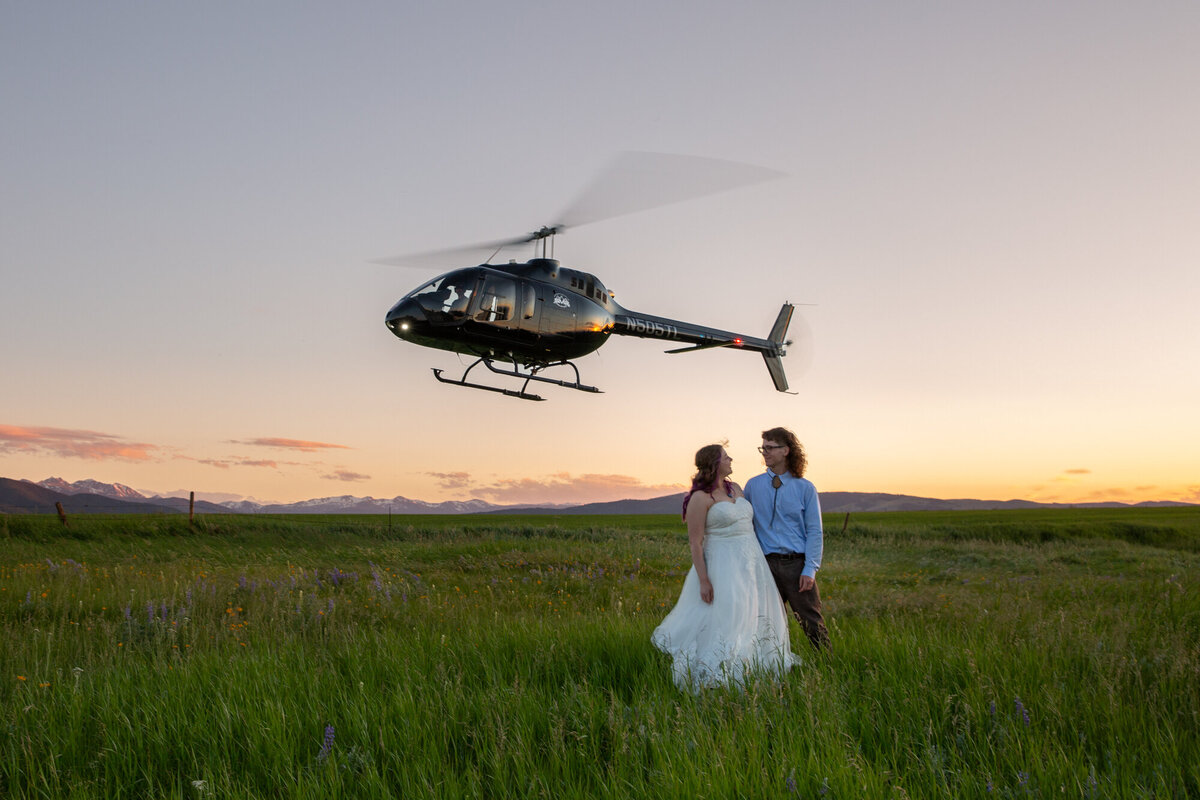 A bride and groom stand in a grassy meadow as a helicopter takes off into the sky behind them.