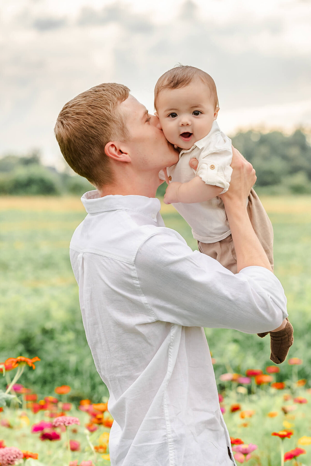 A man wearing a white shirt holds up his infant son and gives him a kiss on the cheek. Image by Justine Renee Photography near the Outer Banks.