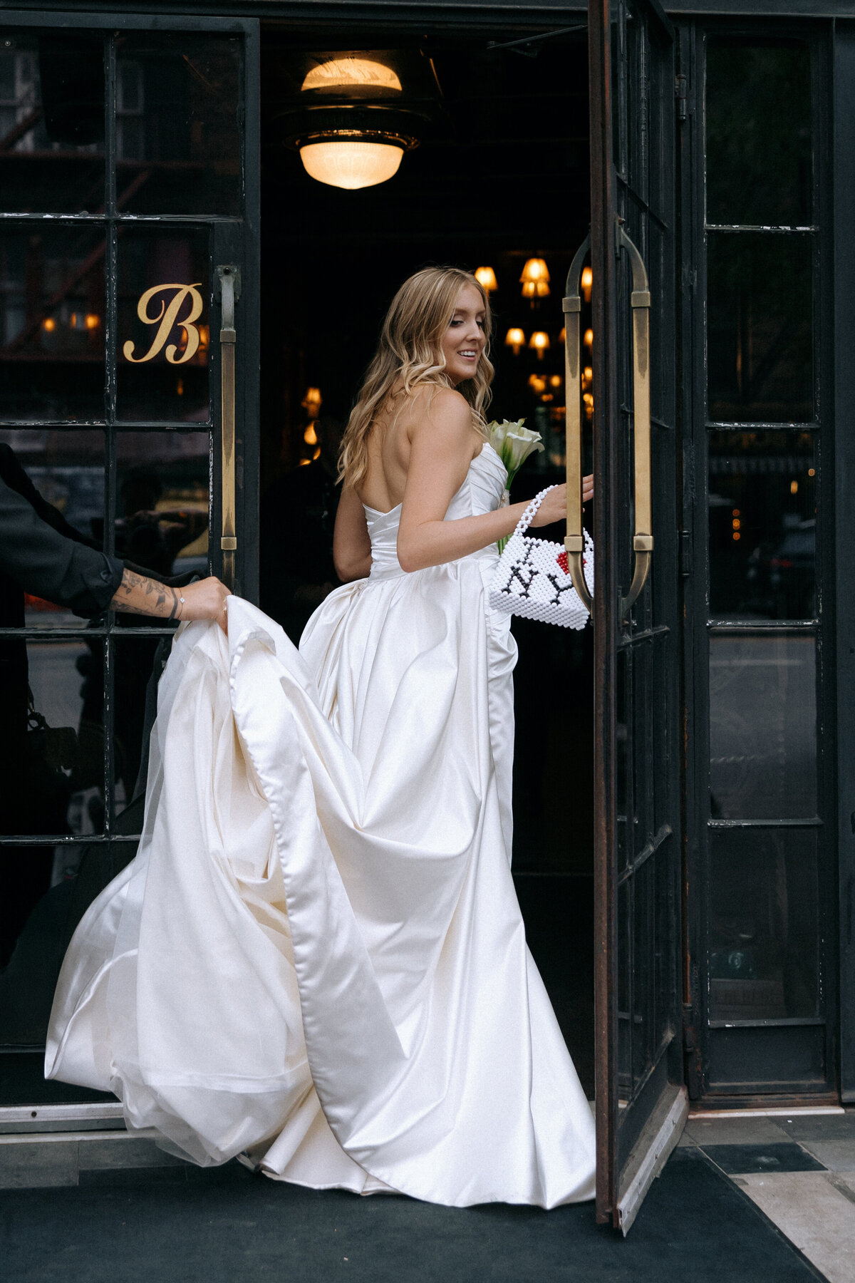 Bride in satin wedding gown entering the Bowery Hotel in New York City, smiling over her shoulder and holding an I ❤️ NY purse, showcasing a modern and fun NYC bridal look