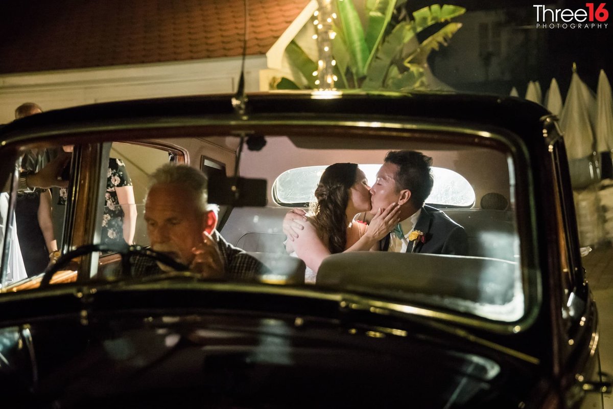 Like an old movie scene, Bride and Groom kiss in the backseat of a car while the driver waits