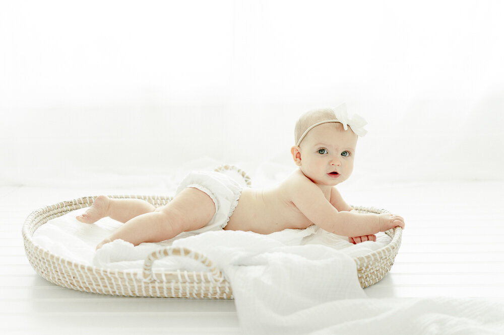 6 month old baby girl lays in a moses basket with a bright window behind her in Kristie Lloyd's nashville photography studio