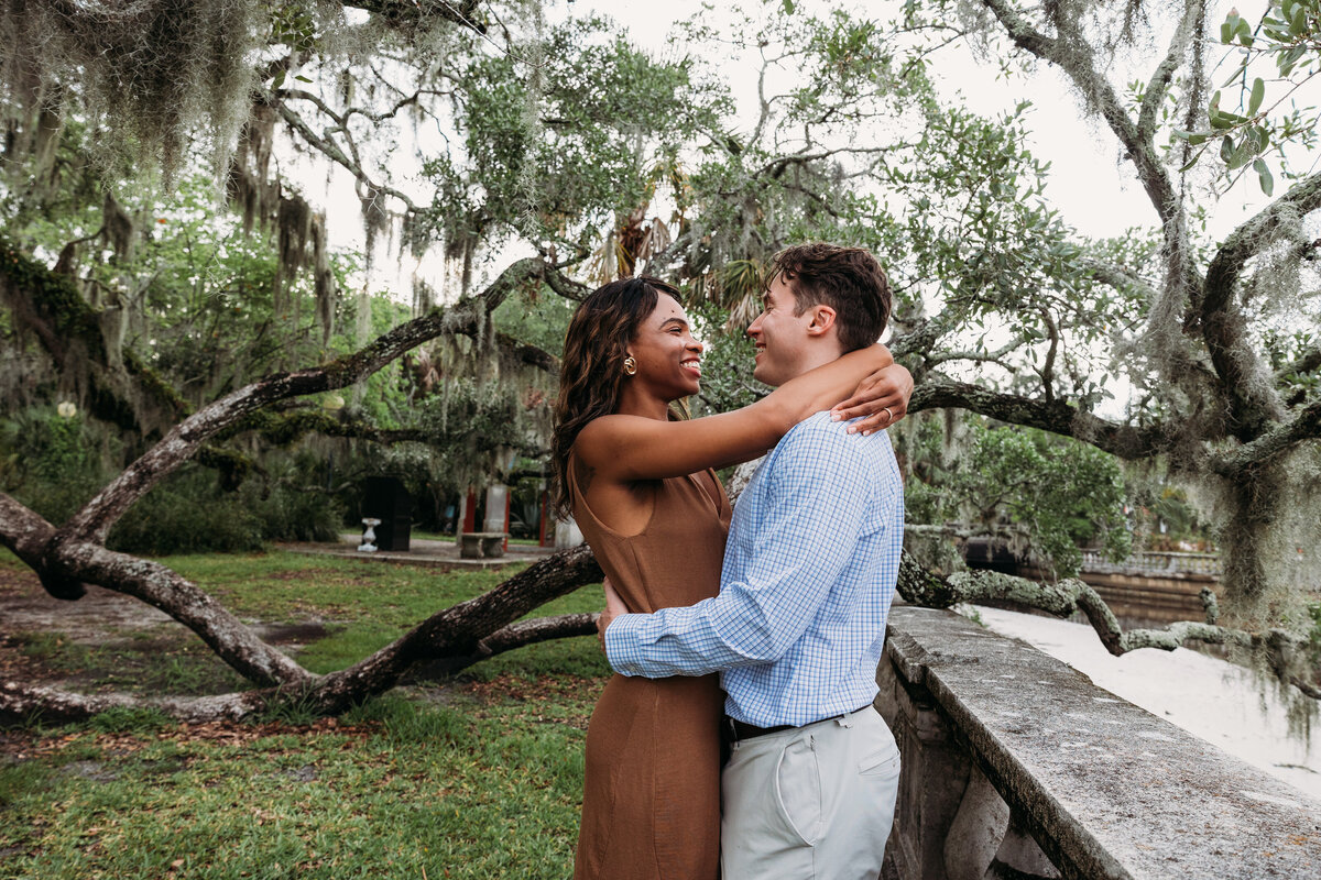 Groom laughs while his fiancée holds his face lovingly during their joyful engagement session beneath twisted tree branches