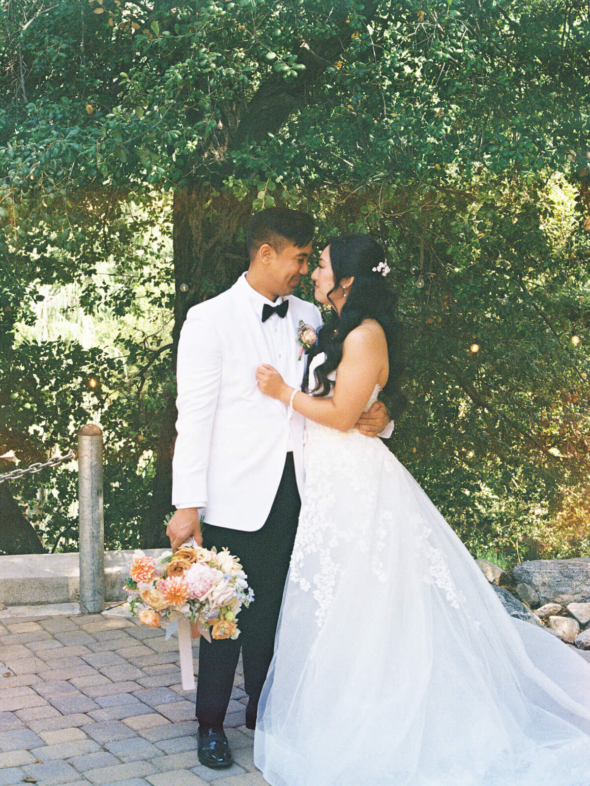 A bride and groom lovingly embrace outdoors. The groom wears a white jacket and holds a bouquet, while the bride wears a lace gown. Sunlight filters through trees.