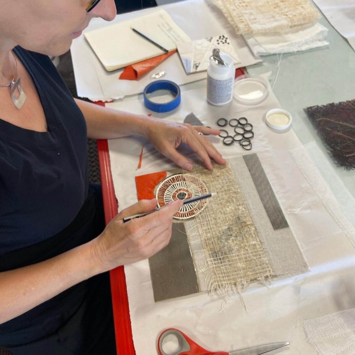 Artist assembling a collagraph plate by arranging textured fabrics and cut shapes, painting adhesive onto a circular patterned element.