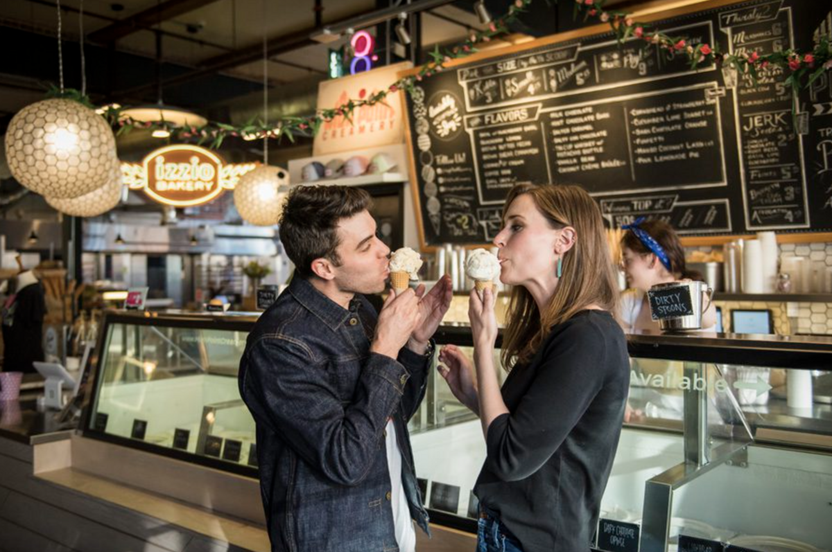 A man and woman stand inside an ice cream shop and both lick a cone with a scoop of vanilla.