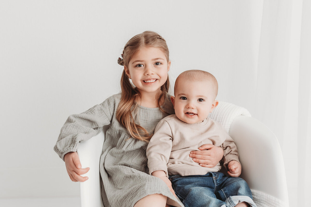 brother and sister sitting in white chair in white photo studio
