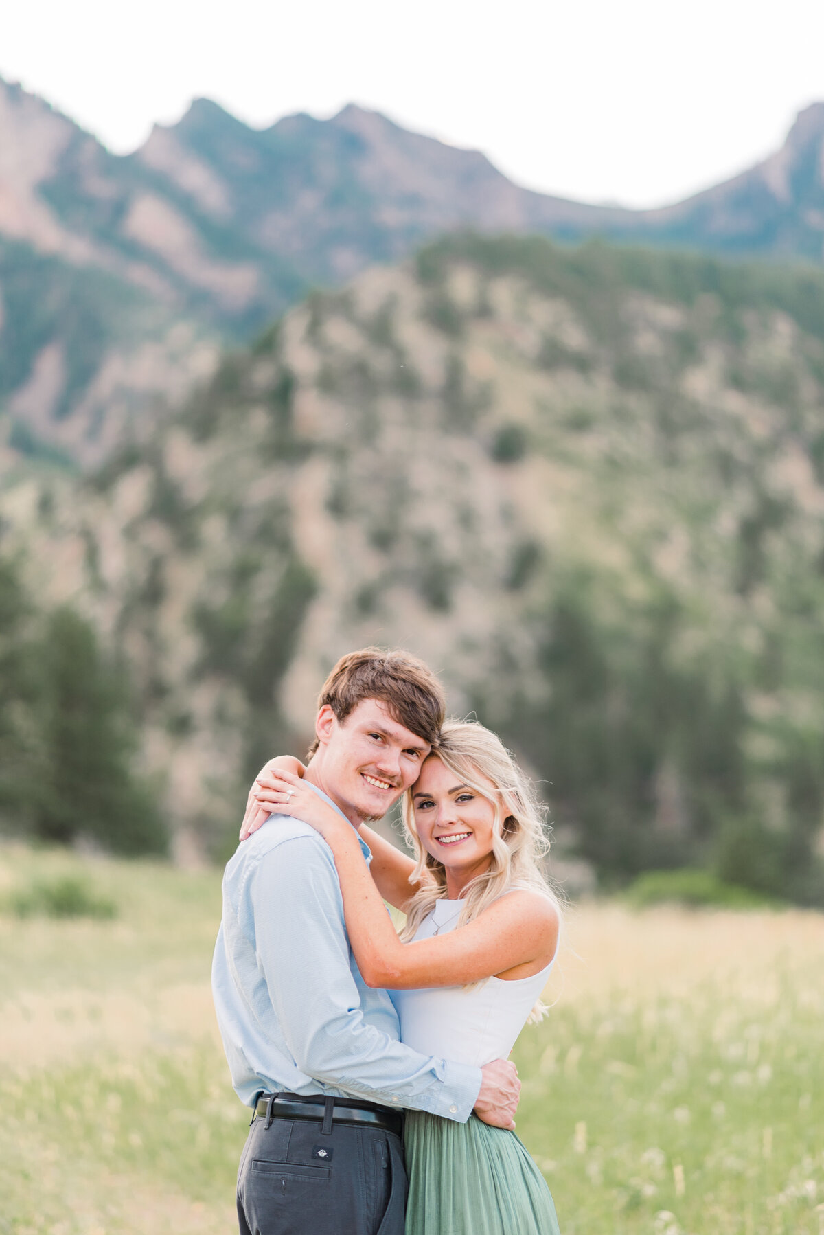 denver engagement photographer captures man and woman embracing in the Rockies with the mans arms around the womans waist as they both smile for their denver couples photography