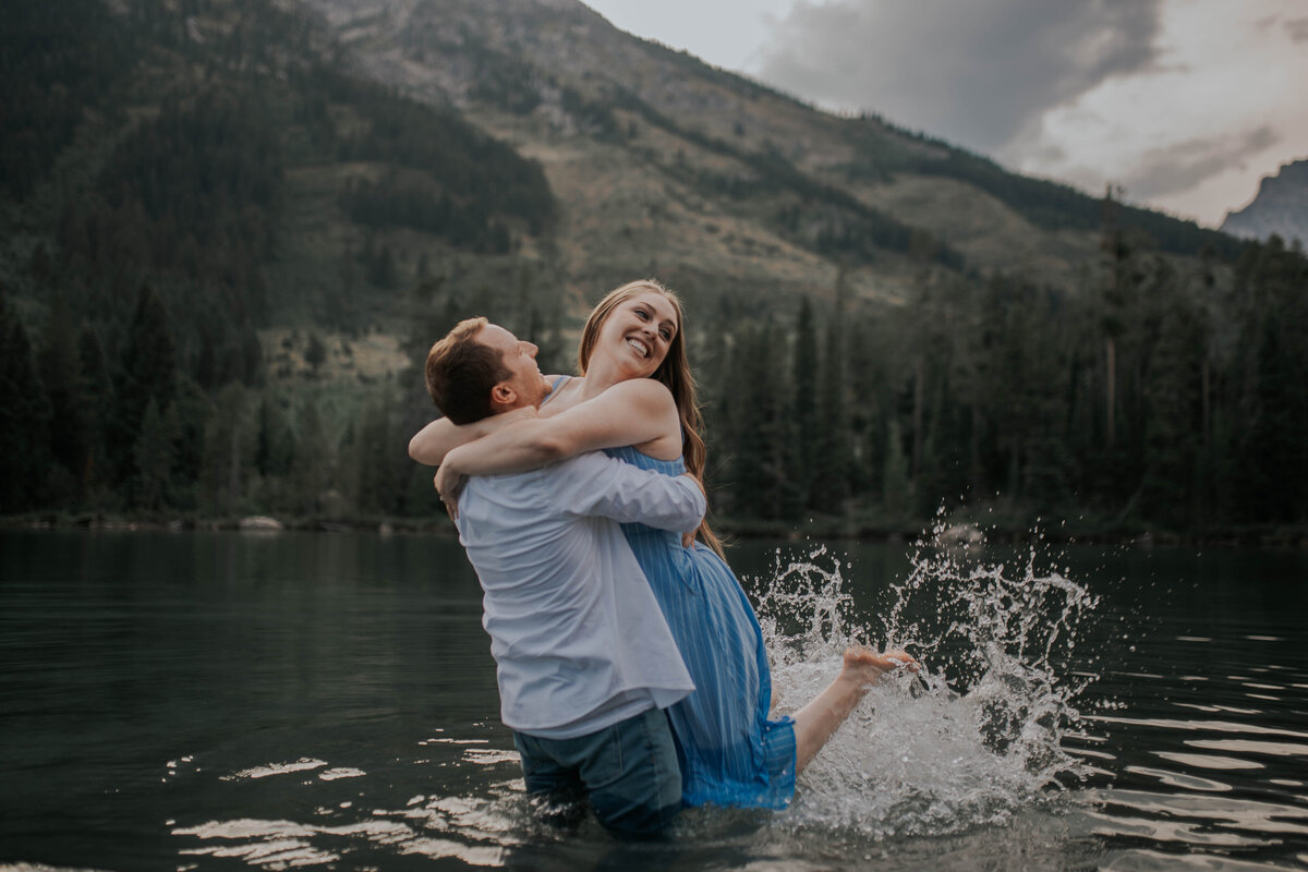 adventurous engagement photos with man and woman playing in the water with the mountains and woods in the distance on the other side of the lake