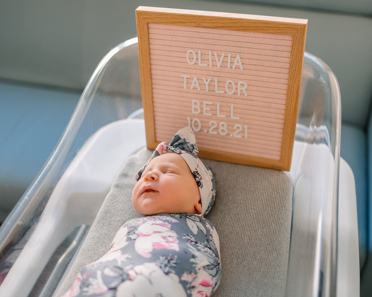 Newborn photograph in a crib with a blanket and a flower headband, above the head is a light pink welcome sign