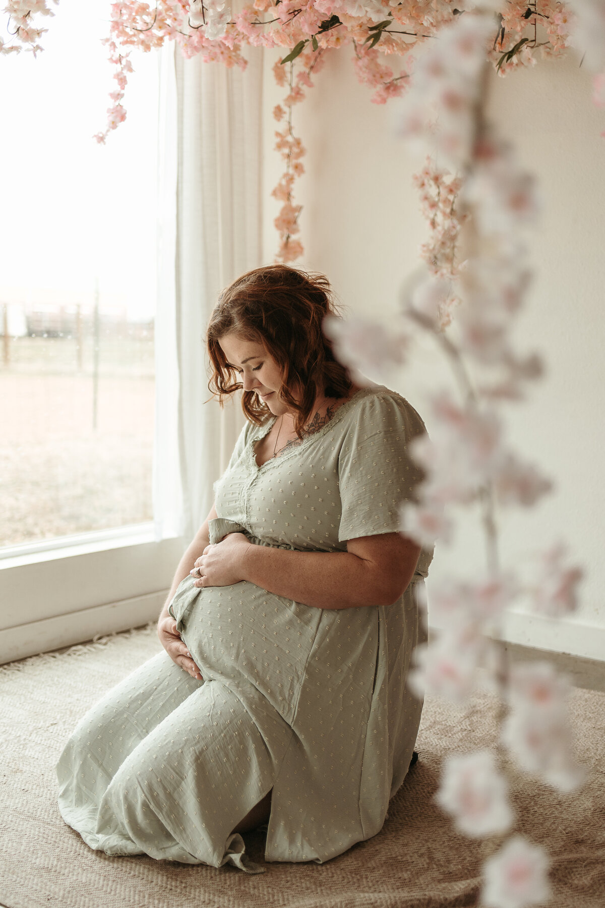 expectant twin mom holding belly and looking down with pink flowers all around her during maternity session with alexis adkins photography in longmont colorado