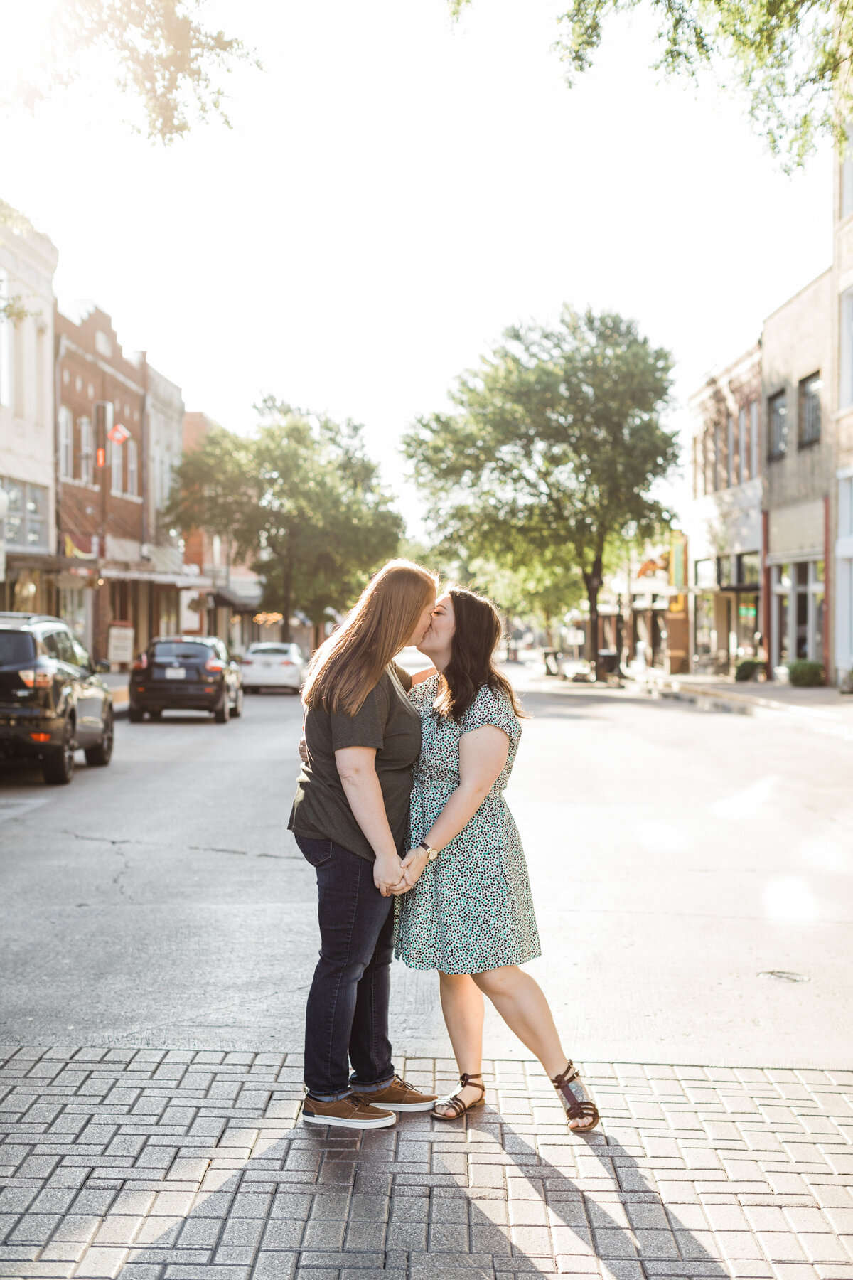 A lesbian couple sharing a kiss during their outdoor engagement session in downtown McKinney, Texas. The woman on the right is wearing a short colorful dress while the woman on the left is wearing a dark t-shirt and dark jeans.