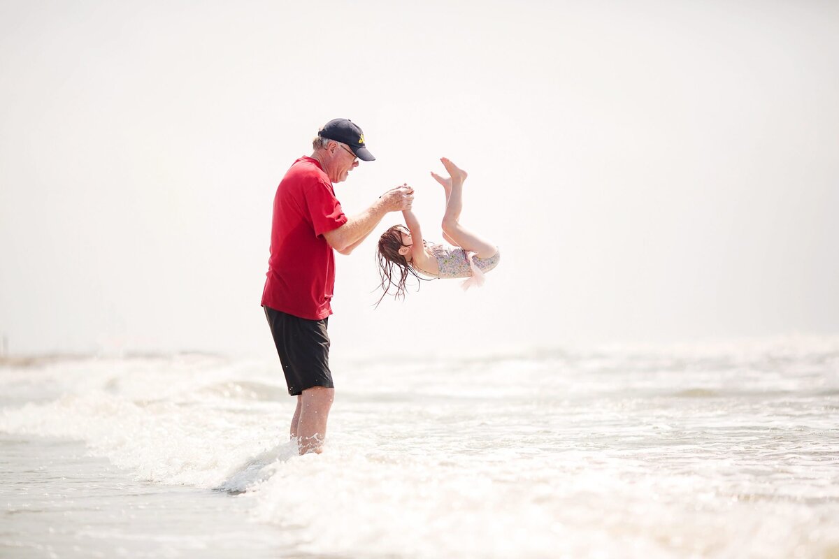 A grandfather in a red shirt plays with his grandaughter at the beach.