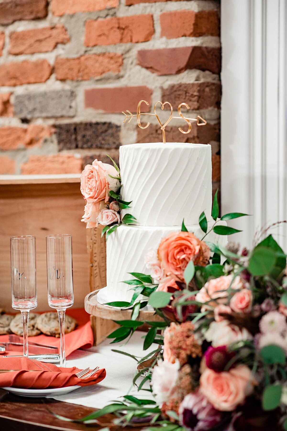 A textured two tier white wedding cake sits in front of an exposed brick wall next to a pair of stemmed champagne flutes with rust napkins. In the foreground is the bridal bouquet of peach roses, blush roses, burgundy dahlias, protea and Italian ruscus with eucalyptus.