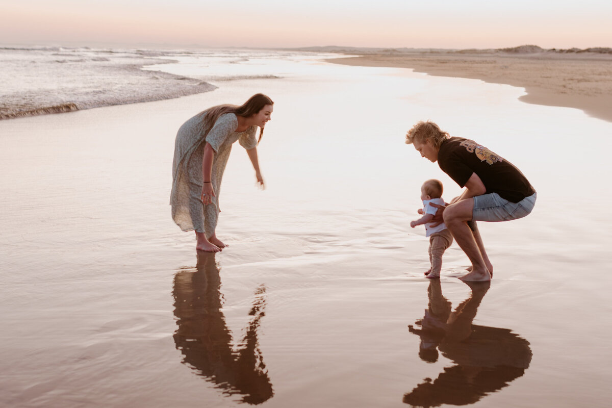 Young family playing at the shoreline during sunset, with parents supporting their baby’s first steps on the wet sand during a relaxed beach photography session.