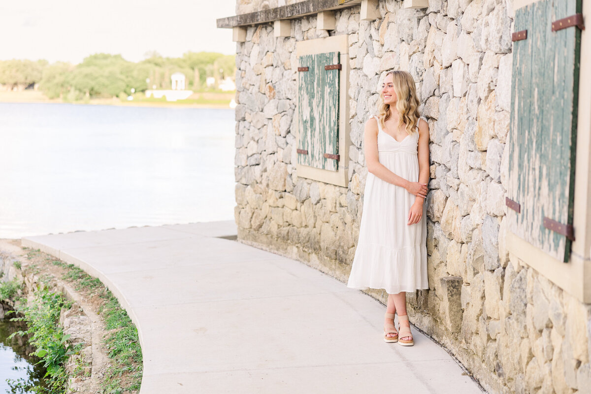 Natural light senior photo of a young woman in a white dress leaning against a stone wall at Adriatica Village in McKinney, Texas. Captured by a McKinney senior photographer specializing in soft, timeless portraits across the Dallas Fort Worth area.