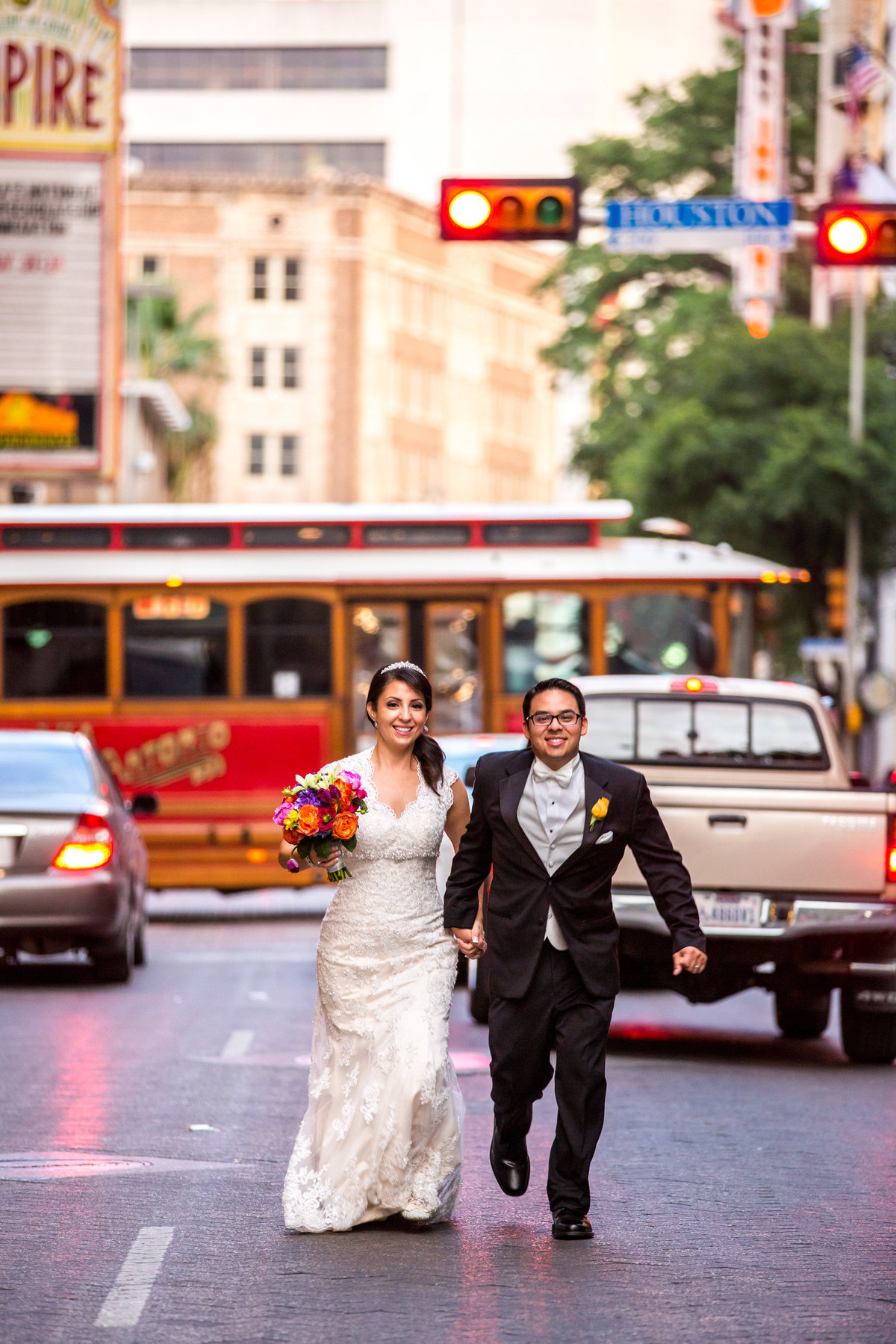 bride and groom running down the street in front of red trolly in downtown San Antonio by the Sheraton Gunter Hotel