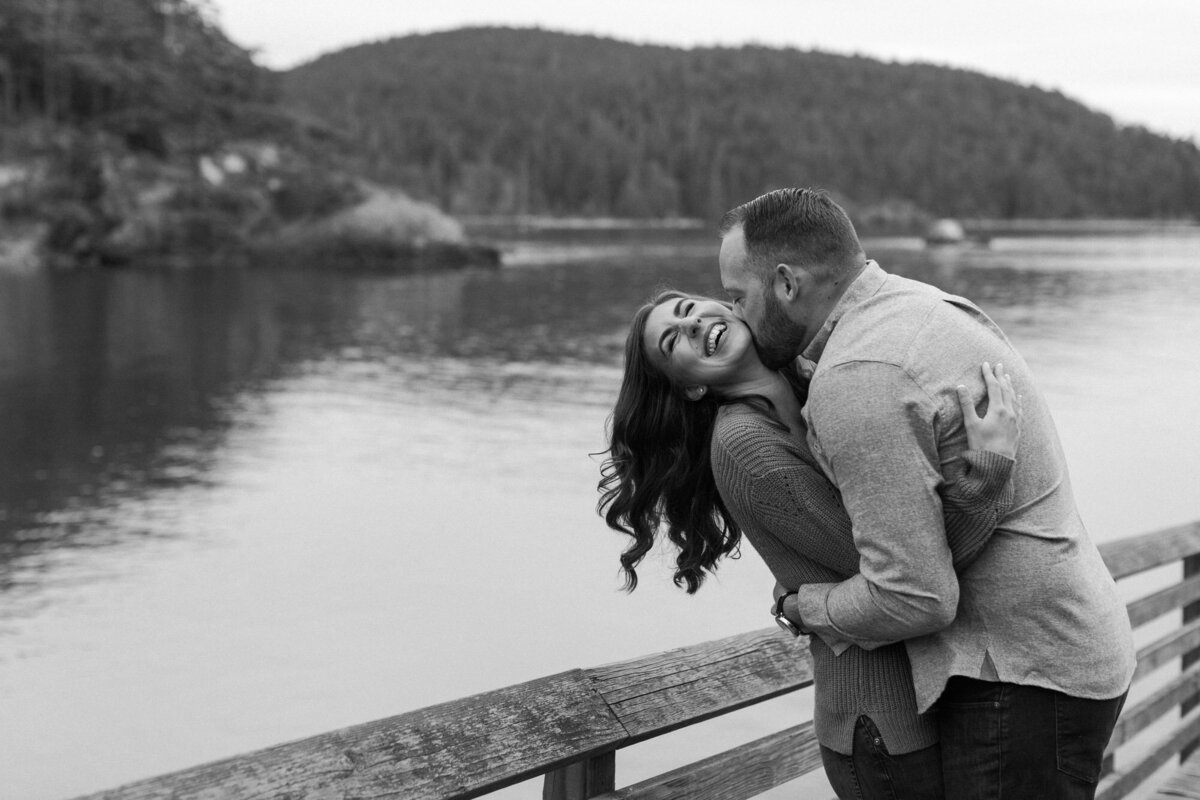 Lauhing couple kiss on dock at Puget Sound near Seattle WA by deception pass black and white candid photo by Joanna Monger Photography