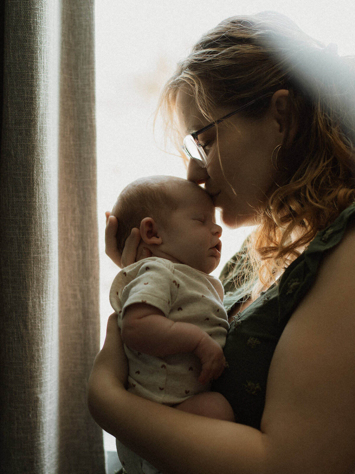 Soft light streams through a nursery window as a mother holds her newborn and kisses her forehead during a cozy in-home session in Lake Elsinore, CA.