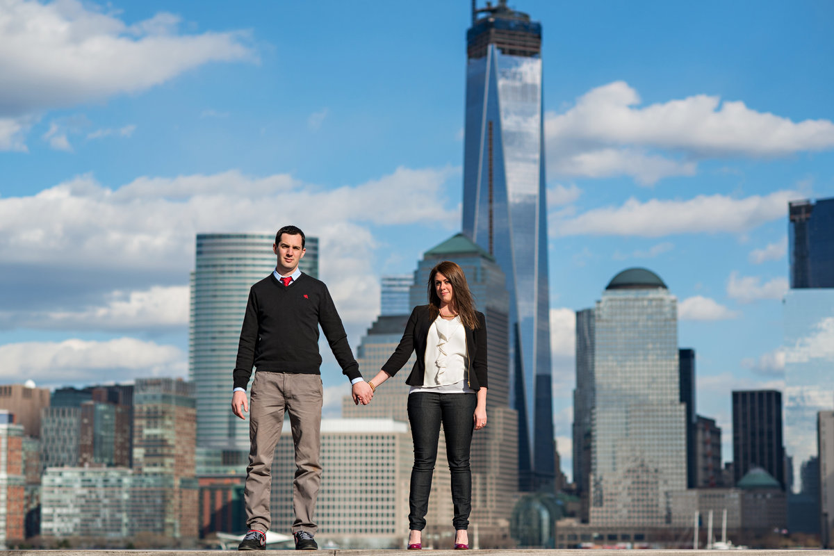 An engaged couple hold hands with the NYC skyline behind them.