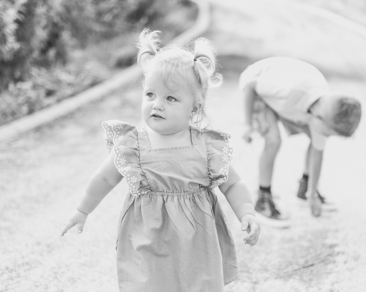 black and white photo of a toddler girl with her older brother in the background on a rocked path.  Toddler is wearing a dress, walking and looking away from the camera while brother is picking up a rock in the background