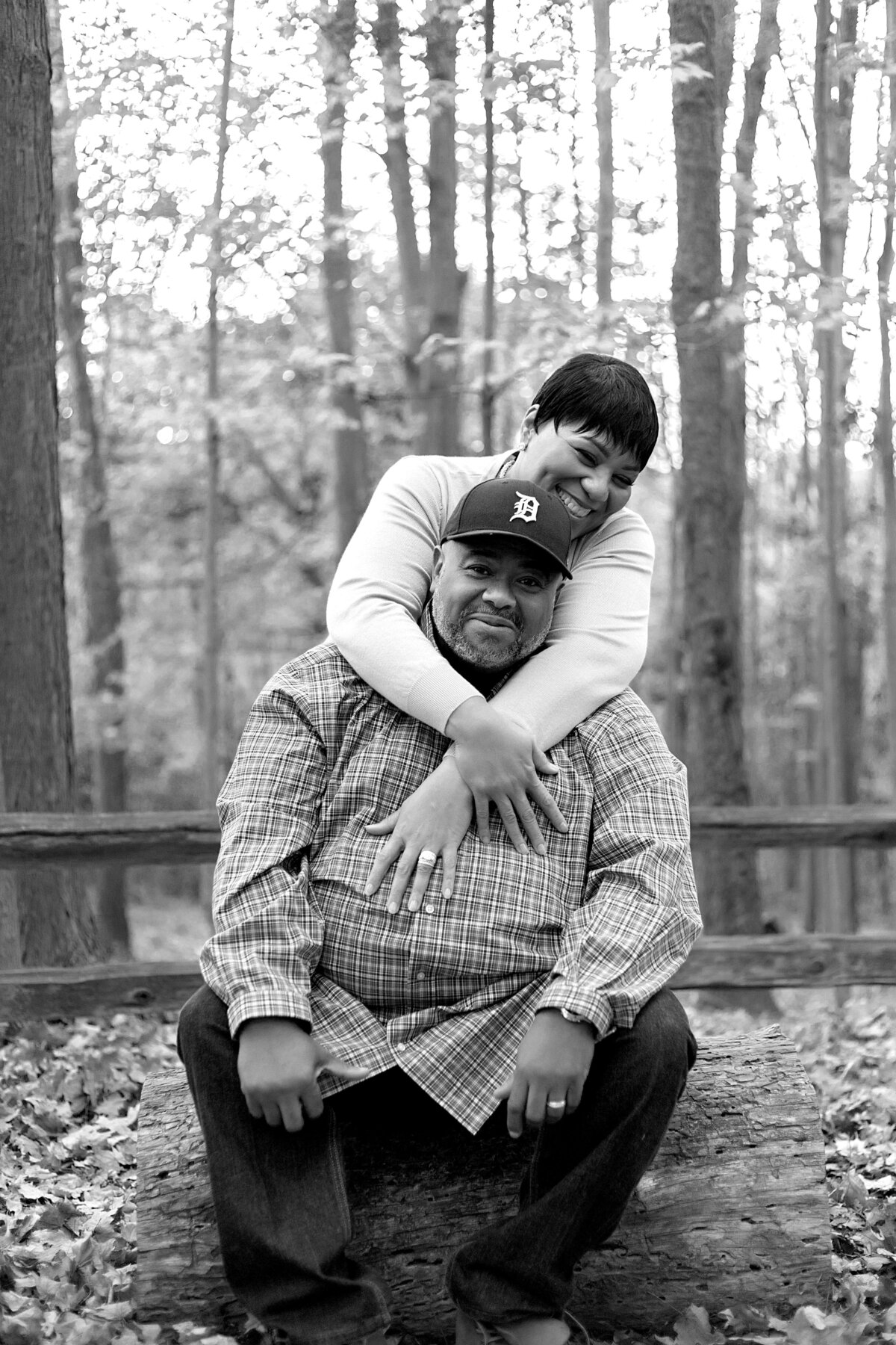Husband sitting on wooden log and wife has arms wrapped around her husband in a park
