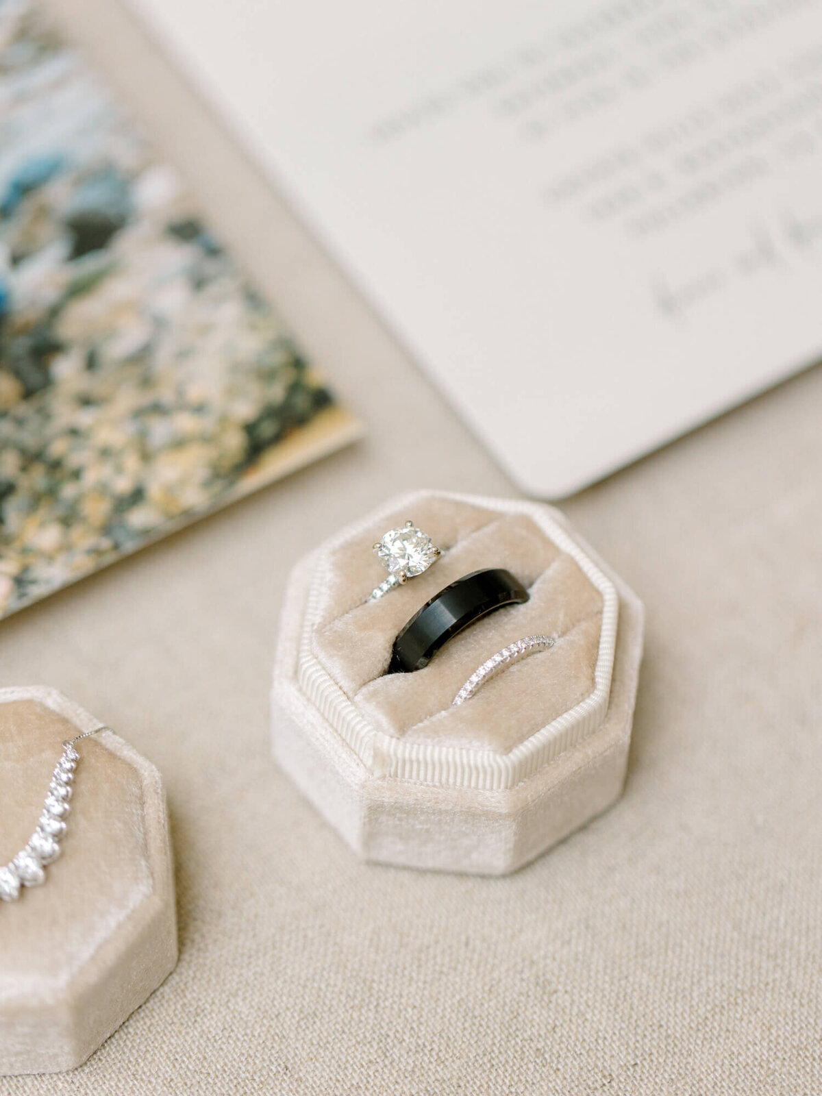 Three rings in a beige velvet box: a diamond ring, a black band, and a thin band with small gems. Background includes a blurred photo and paper.