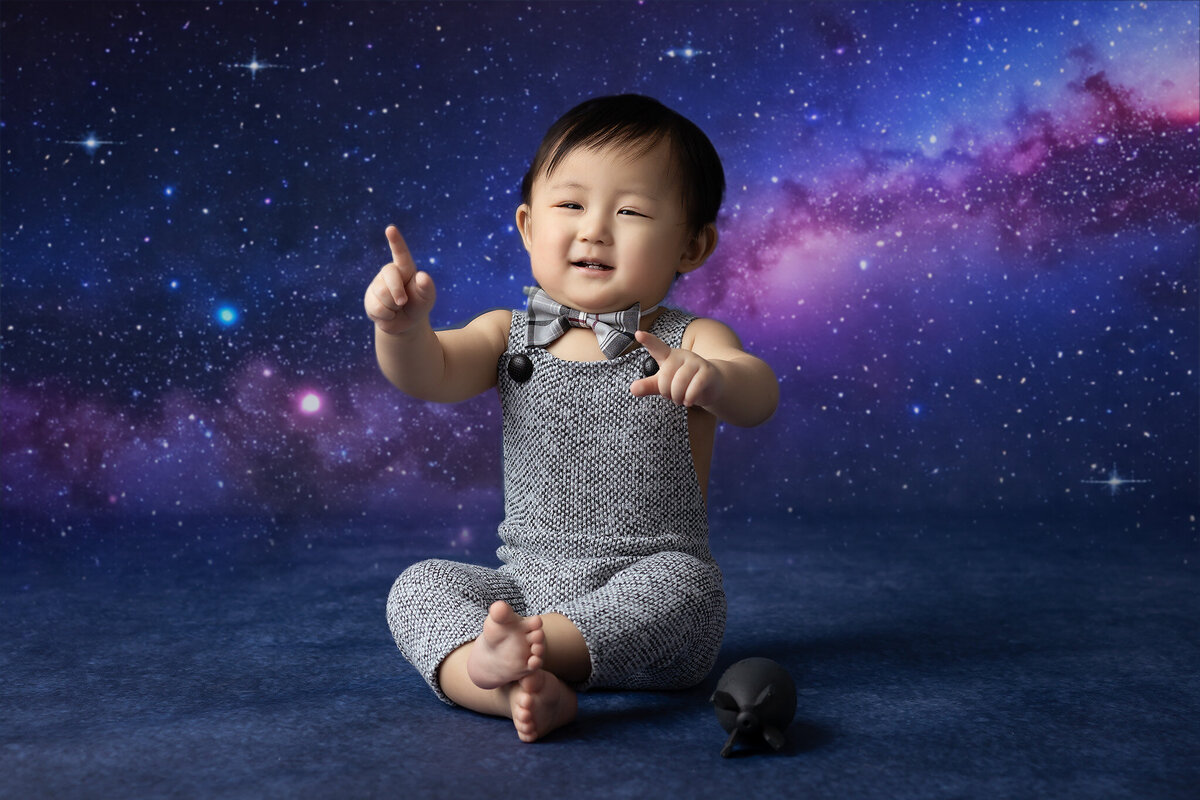 A baby boy in a bowtie and onesie points at a starry galaxy backdrop, sitting with a toy.