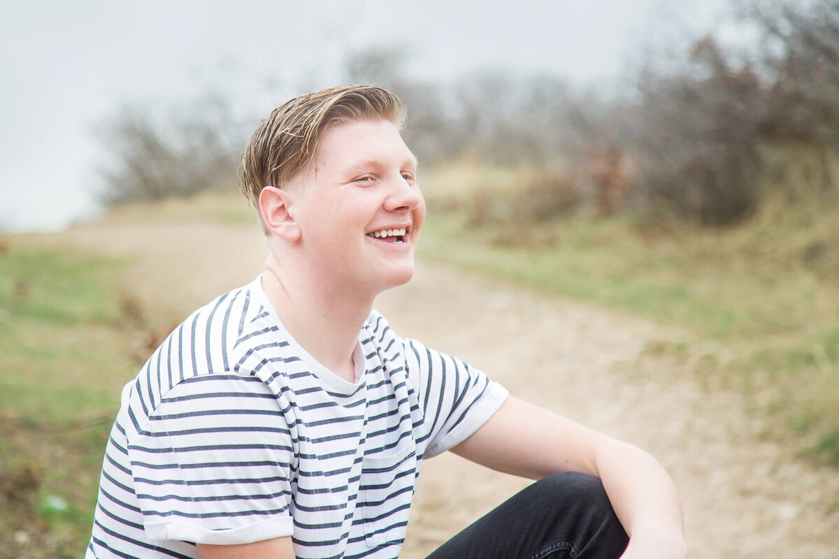 Young man in a black and white striped t shirt laughing while sitting on a rock during his session with las vegas milestone photographer