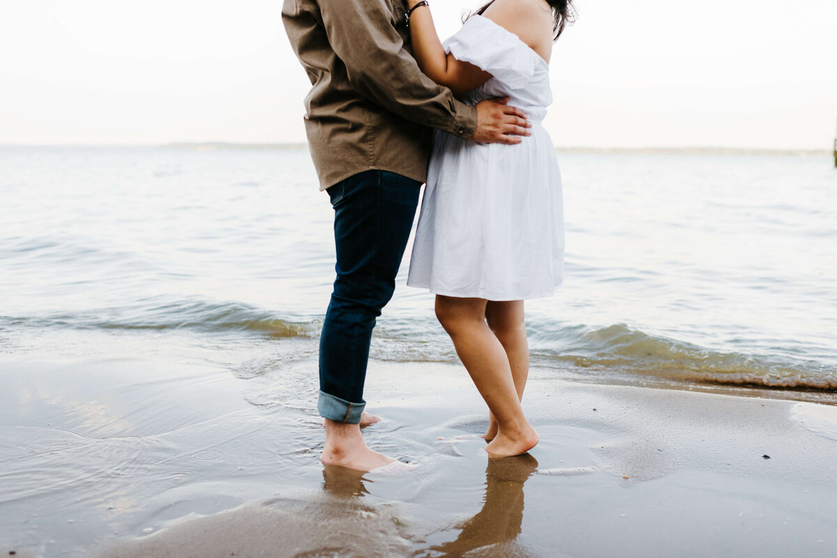 woman in a white dress standing with a man in jeans on the shore of woodbridge lake for beach engagement photos as they embrace together in the water