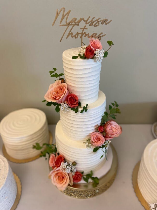 A table filled with cakes - a three tier wedding cake is surrounded by single tier kitchen cakes. All cakes have white frosting with line texture. The tiered cake also has clusters of flowers in shades of pink alternating sides.