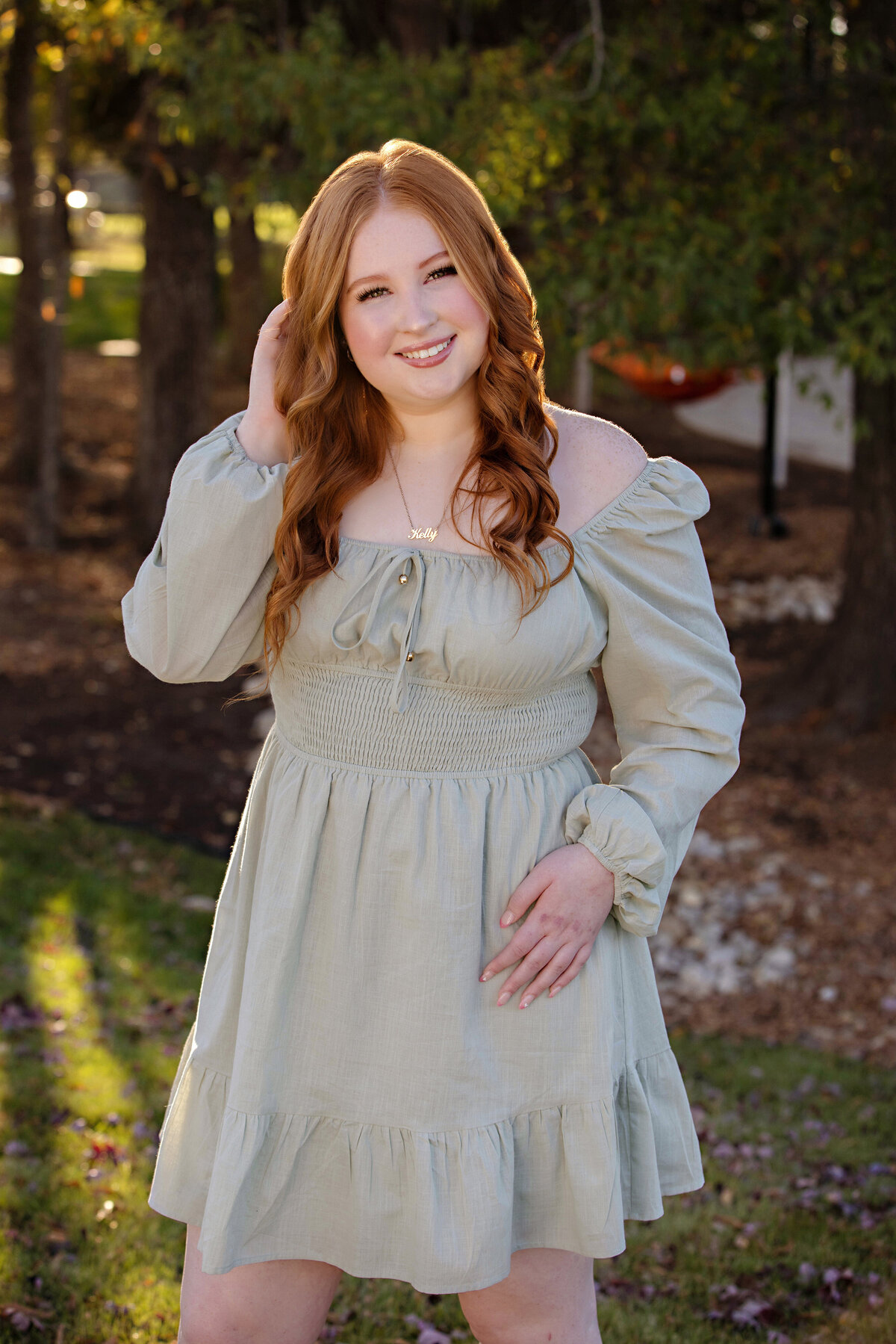 High school senior in sage green dress standing near lush foliage in Ballantyne, sunlight catching her red hair