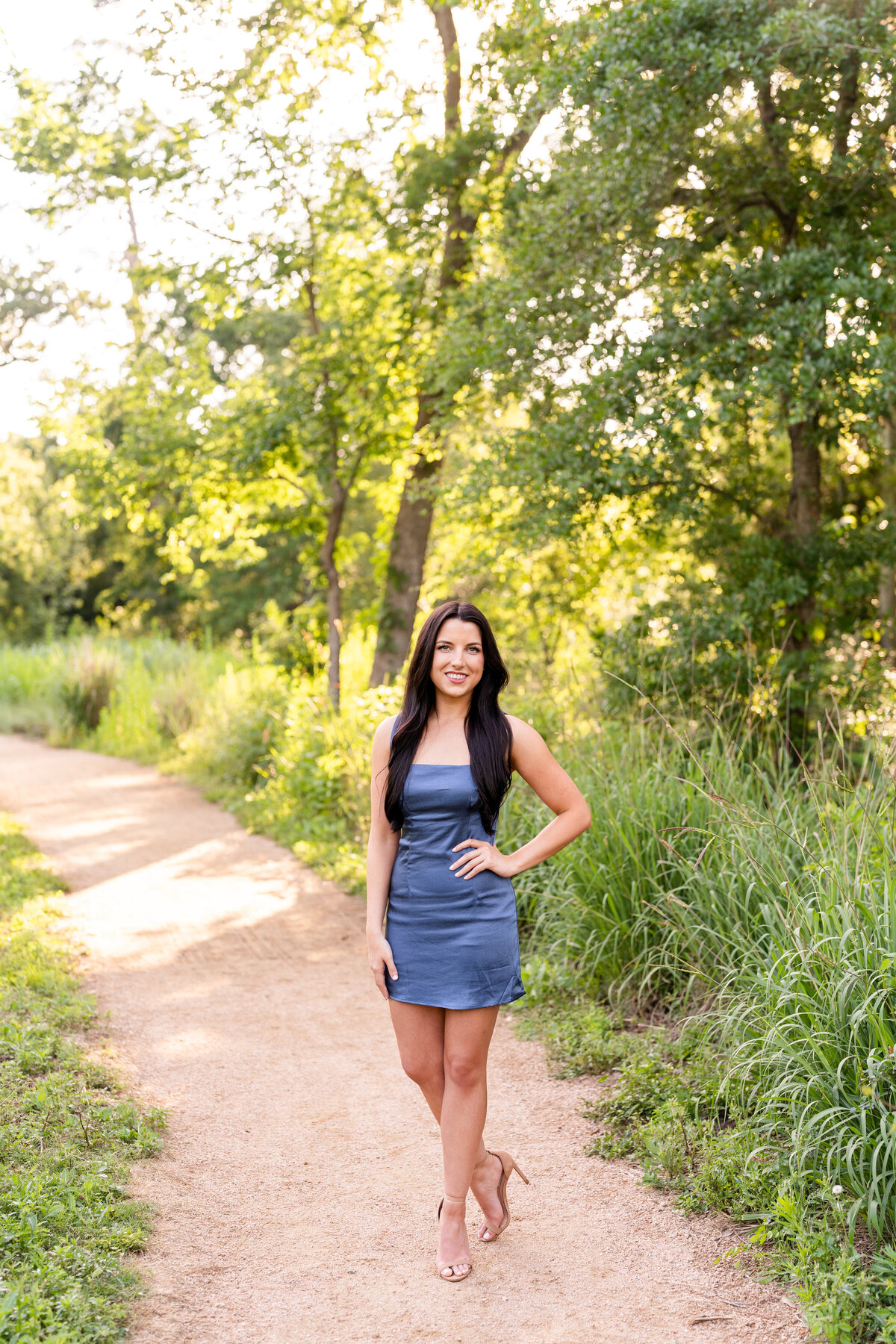 Baylor senior girl standing with hand on hip in blue dress on pathway surrounded by greenery at Houston Arboretum at sunset