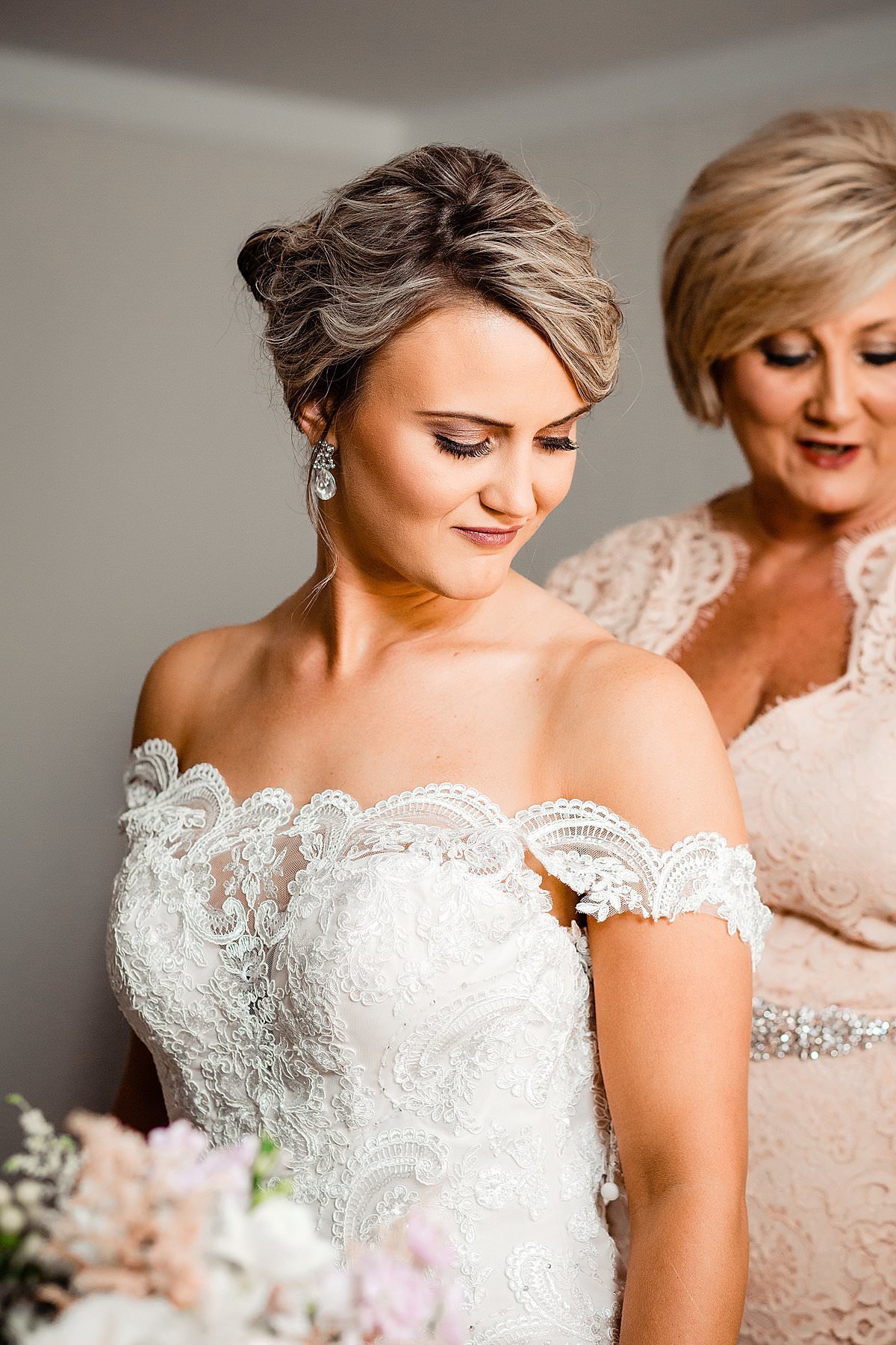Bride wearing a lace dress with her hair up and mom helping zip her into her dress
