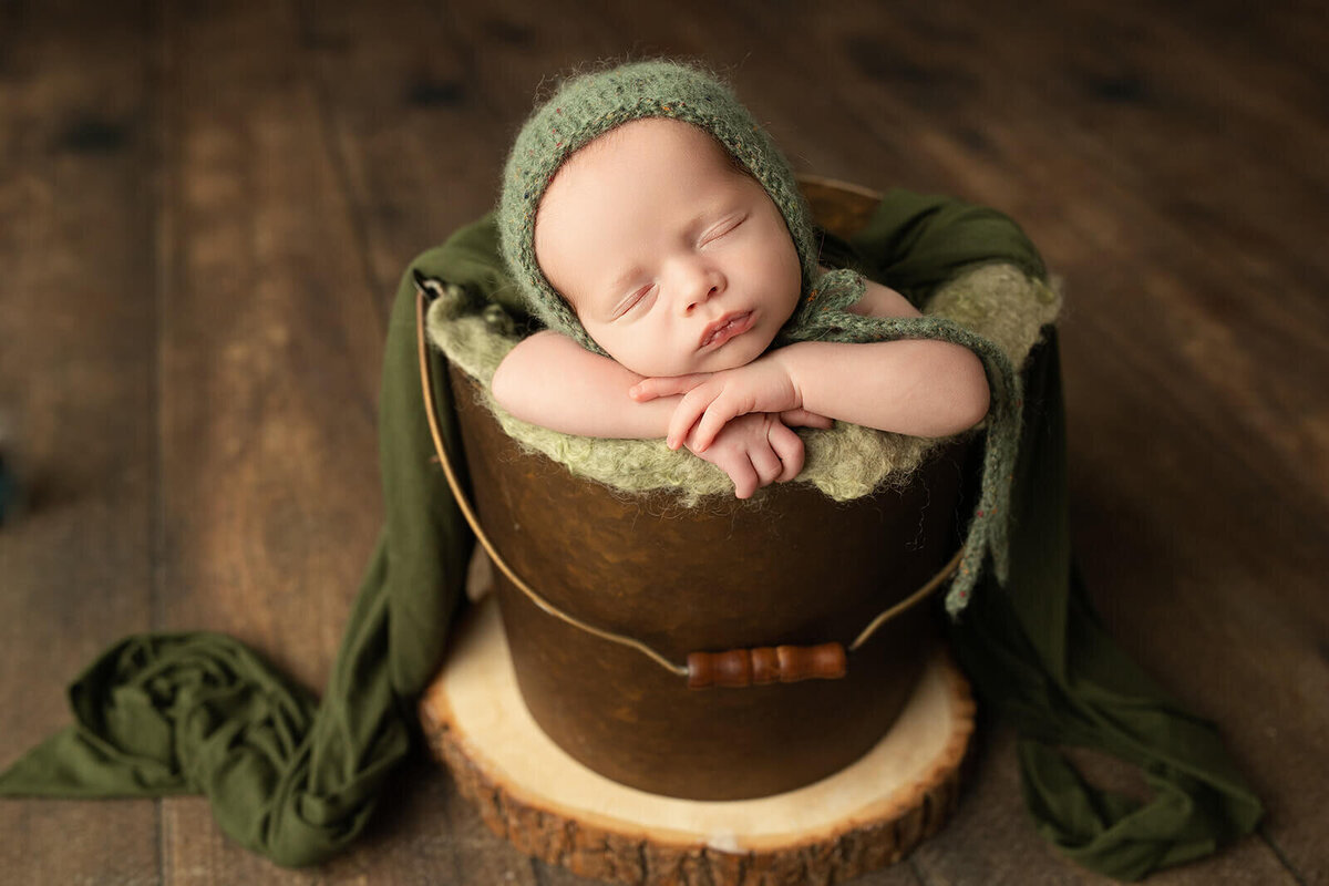 baby in a bucket sleeping with a green bonnet on a slab of wood for his newborn photos