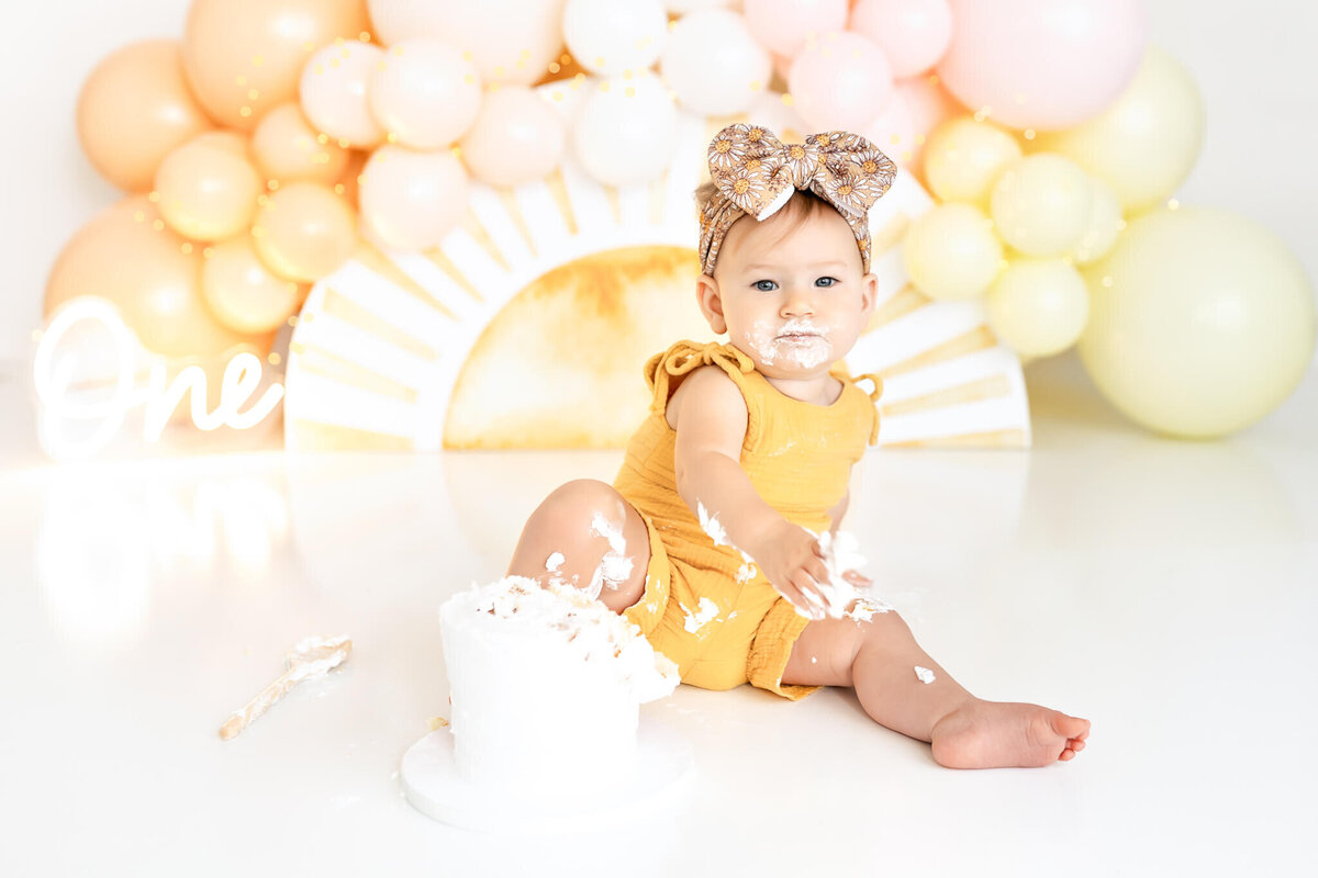 A baby wearing a yellow outfit and a bow sits on a glossy floor surrounded by pastel balloons. There's a partially eaten white cake in front of them, and "One" is displayed in white script in the background.