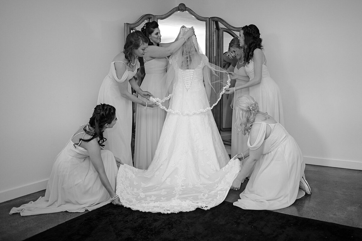 Black and white image of Bride standing in front of antique mirror as bridesmaids adjust veil and wedding gown