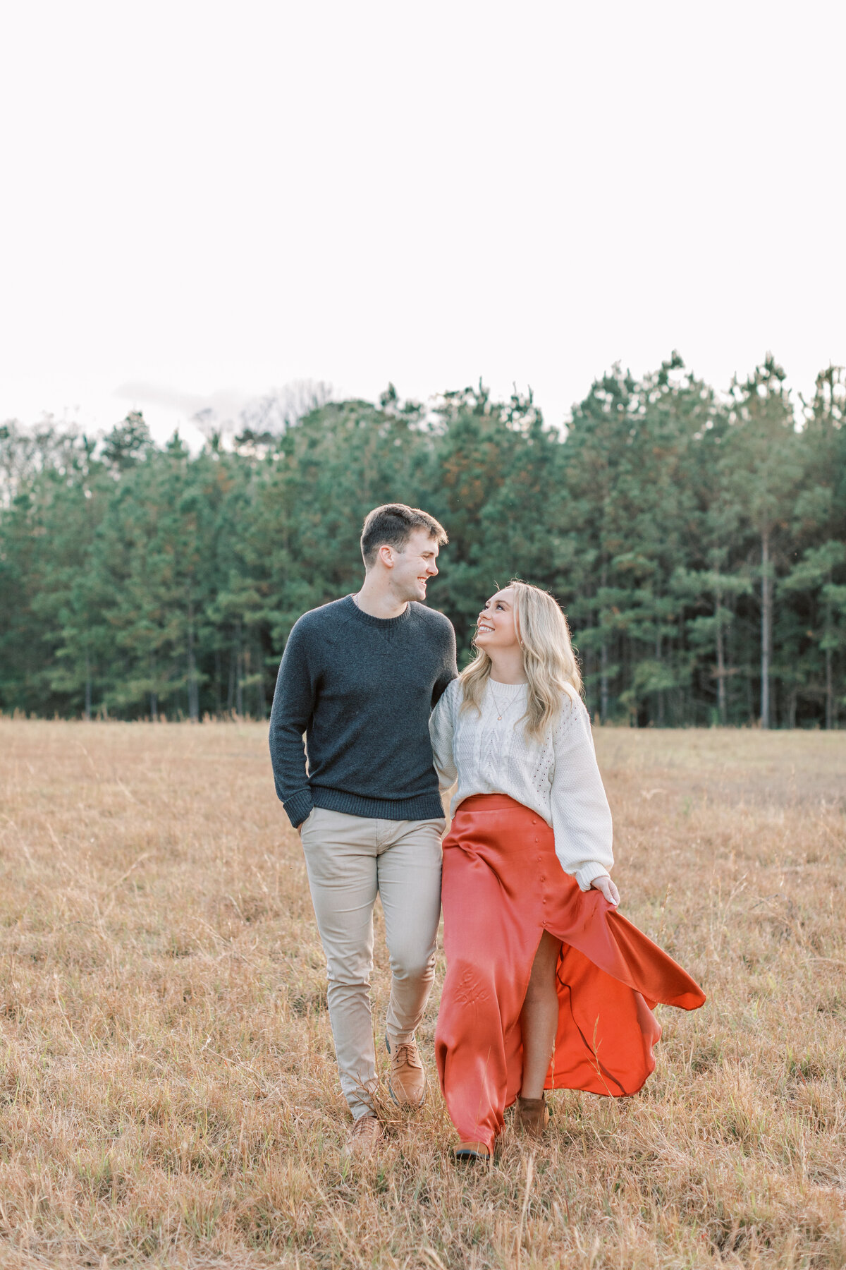 A couple walks through a field surrounded by pine trees.