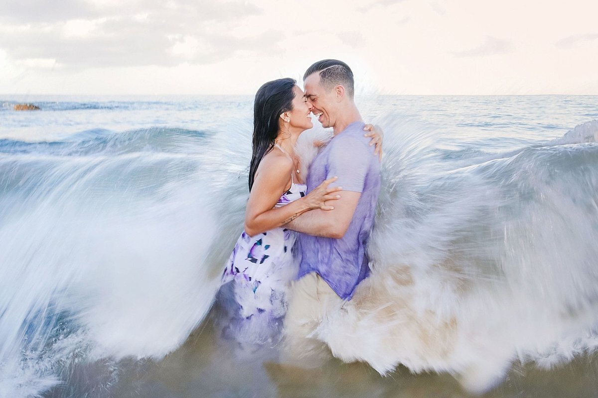 Shorebreak washes over a couple laughing in the ocean while their engagement session is photographed by Love + Water on Maui
