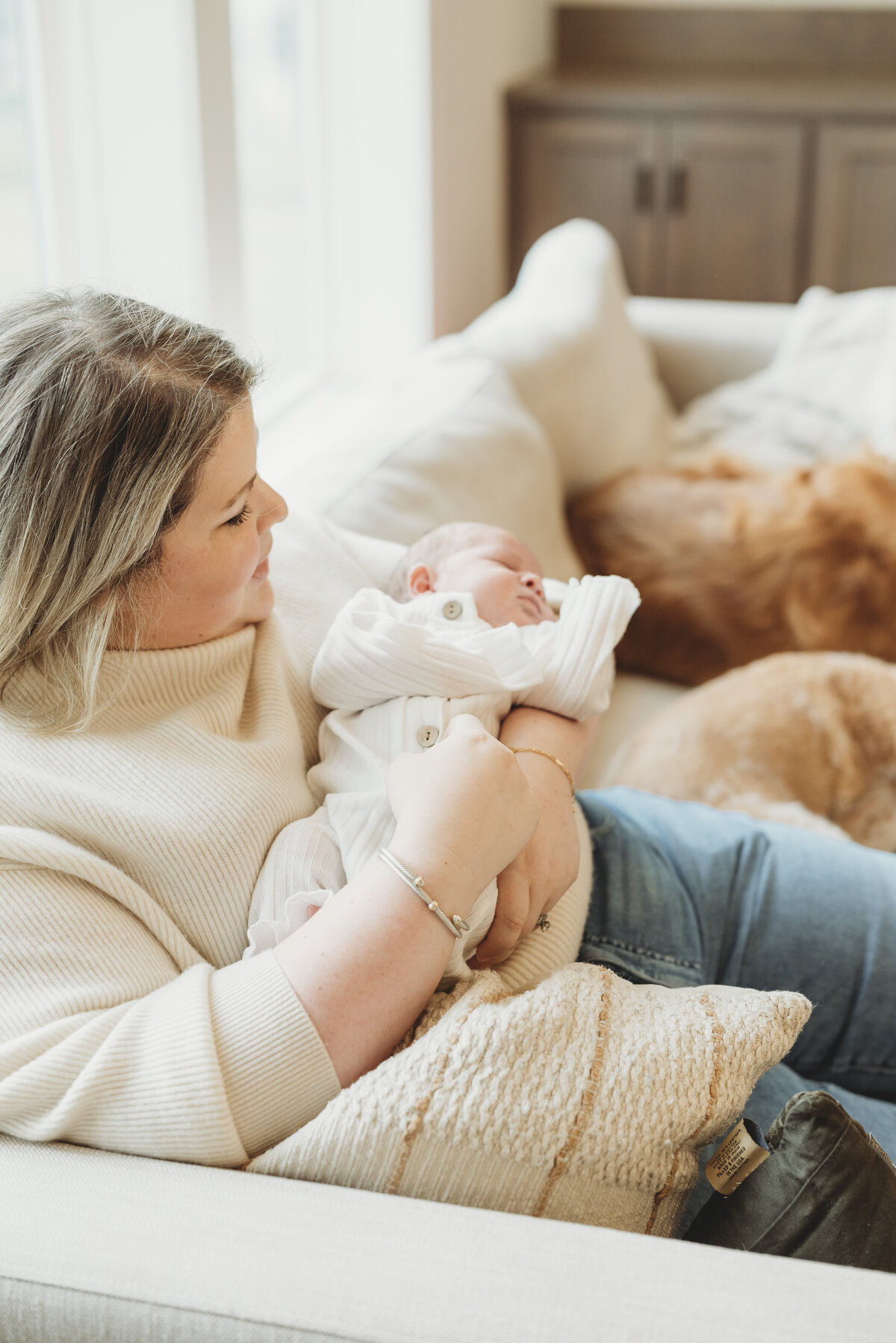 Mother sitting on a sofa, smiling while holding her newborn baby during a cozy in-home Kansas City newborn photography session.