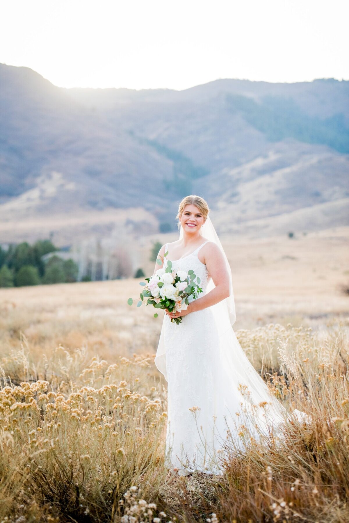 A bride holding her bouquet stands in a golden field and smiles at the camera at her wedding at The Manor House in Littleton, Colorado.