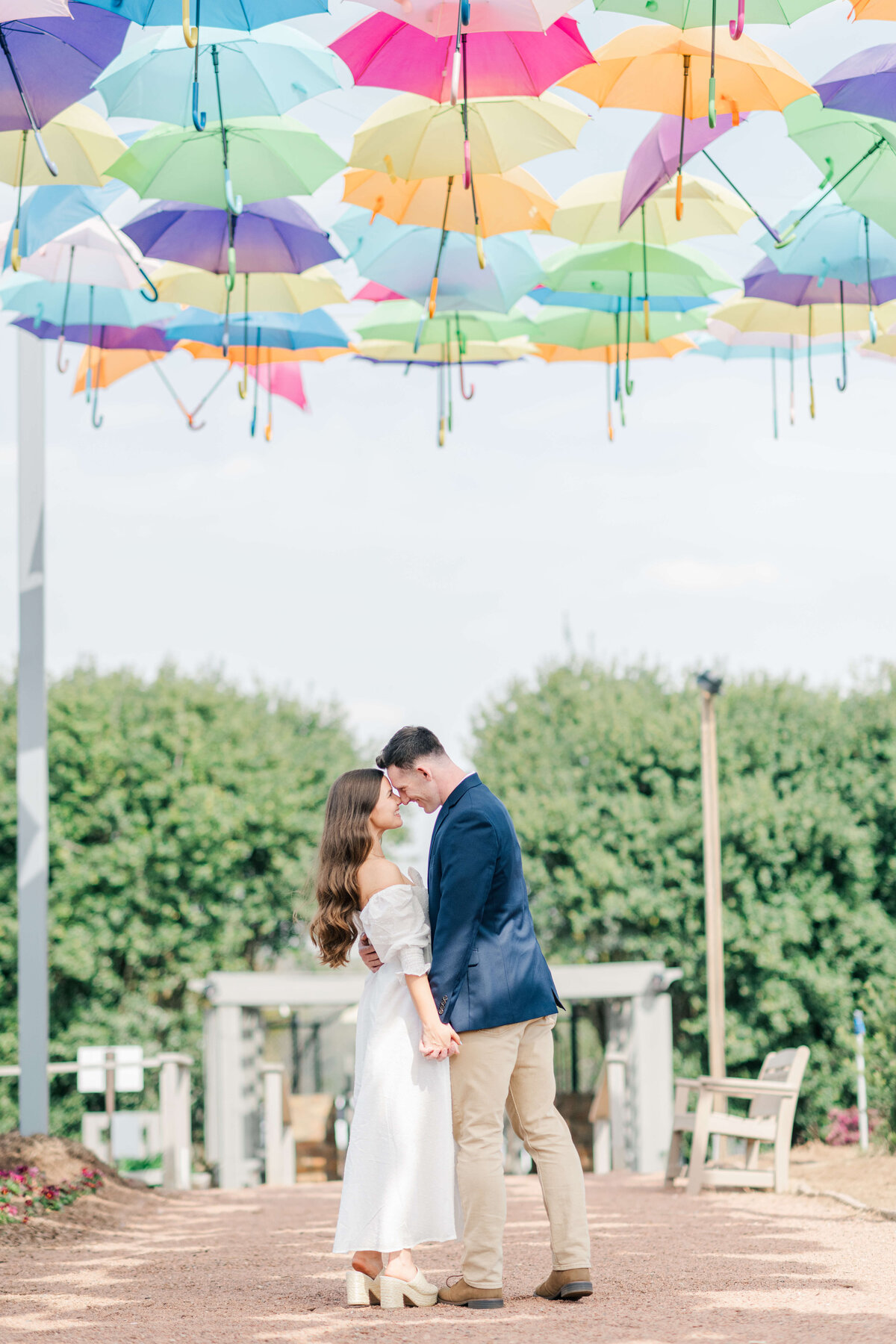 Man and woman stand under colorful umbrellas while looking into each others eyes and leaning towards each other