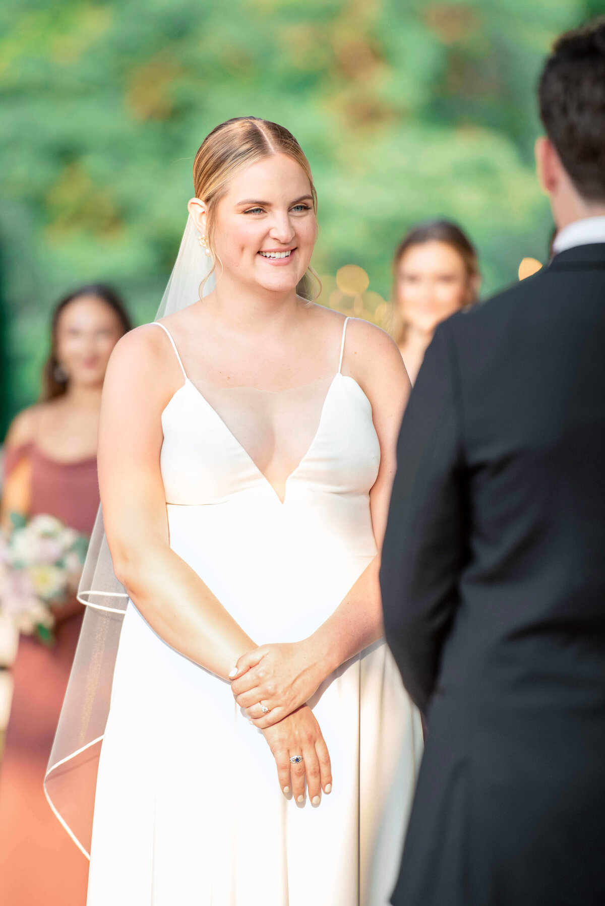 A bride in a white dress and veil stands outdoors, smiling at a groom in a black suit. Blurred bridesmaids in the background hold flowers. The setting is lush and green.