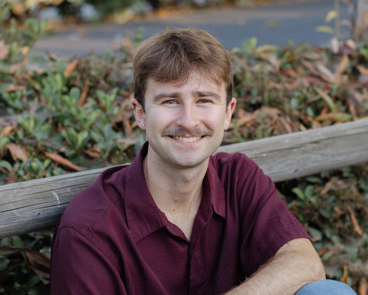 A close-up of a young man with dirty blonde hair and a mustache, wearing a red shirt, leaning against a park fence and smiling up at the camera.