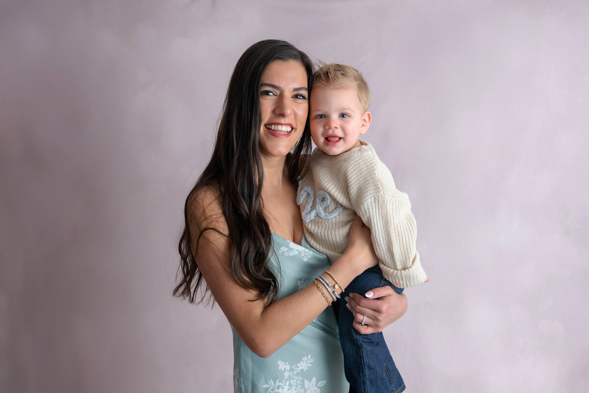 Woman with long dark hair holding a smiling toddler cheek to cheek, both looking forward with candid smiles.