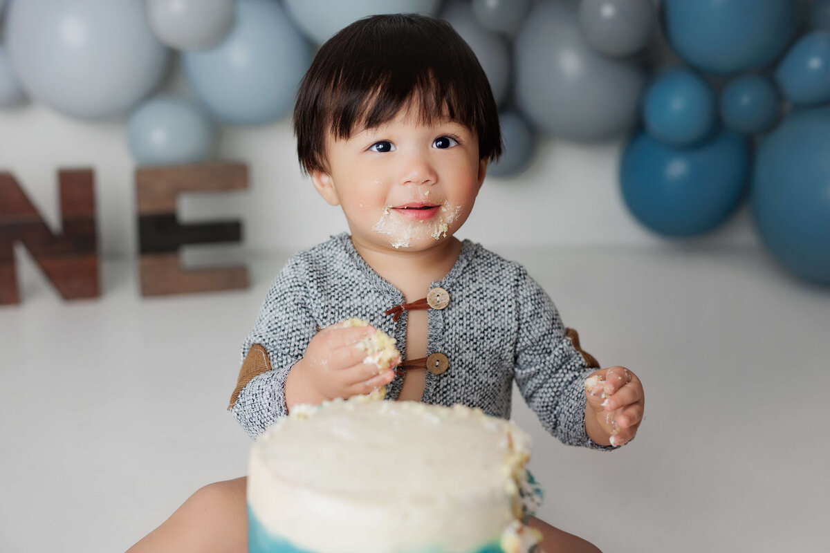 one year old boy sitting in front of a birthday cake with balloons and one letters a t cake smash session