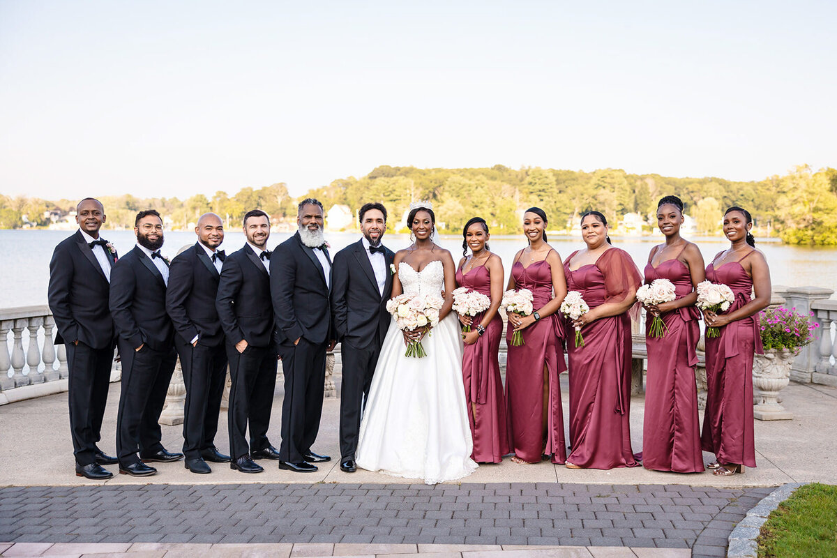 A wedding party portrait by a lake, with the bride and groom centered among groomsmen in black tuxedos and bridesmaids in long burgundy dresses