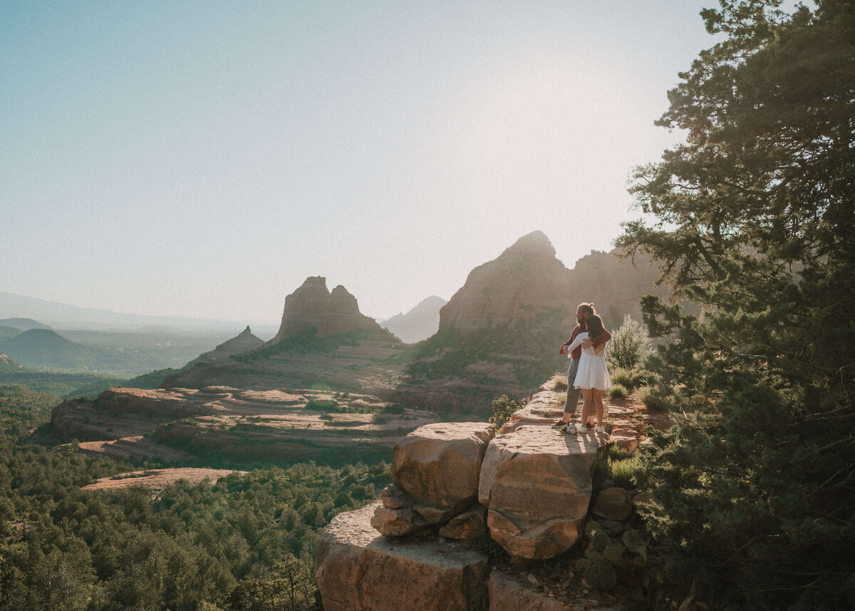 couple embracing on side of mountain range with Sedona landscape behind them