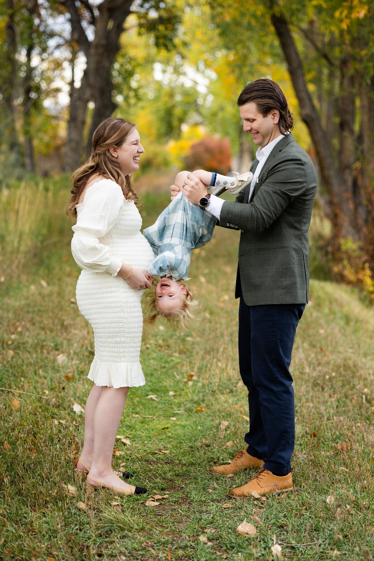 Dad holds toddler daughter upside down while she laughs and hugs mom's pregnant belly.