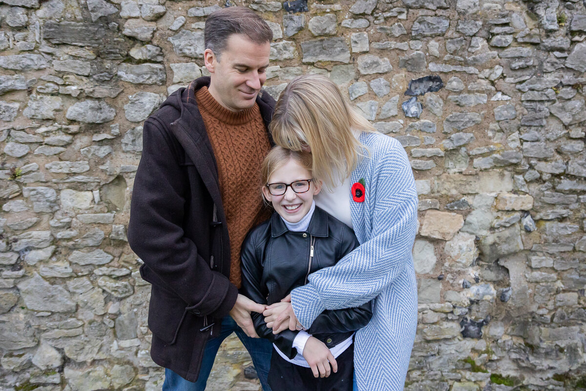 A family posing in front of a stone wall during a photography session, mother is giving a kiss on her head to her daughter