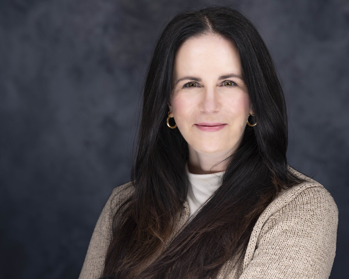 Headshot in CT of woman with long dark hair in beige sweater, smiling against dark textured background by Rob Faber Photography.